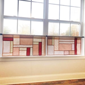May include: Two cafe curtains hanging on a black rod, featuring a geometric patchwork design composed of rectangular fabric panels in shades of white, beige, light pink, and deep red, separated by thin brown lines across the window.