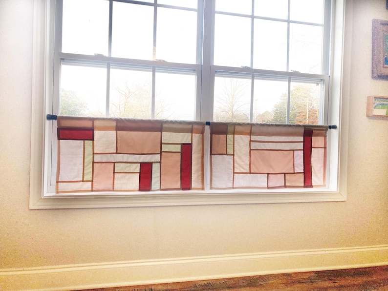May include: Two cafe curtains hanging on a black rod, featuring a geometric patchwork design composed of rectangular fabric panels in shades of white, beige, light pink, and deep red, separated by thin brown lines across the window.