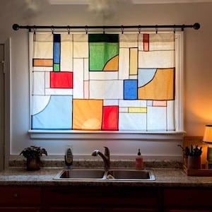 May include: A window curtain featuring a geometric pattern of rectangular and curved shapes in primary colors including red, blue, yellow, and green on a white background. The curtain is hung on a black metal rod with rings over a kitchen sink.