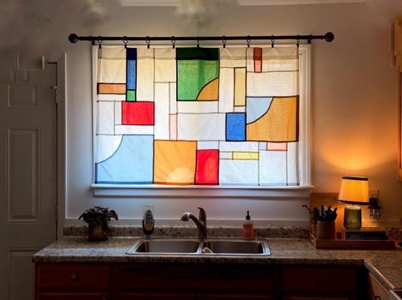 May include: A window curtain featuring a geometric pattern of rectangular and curved shapes in primary colors including red, blue, yellow, and green on a white background. The curtain is hung on a black metal rod with rings over a kitchen sink.