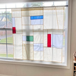 May include: A window curtain featuring a geometric grid pattern on a cream-colored background. The design includes rectangular blocks of solid blue, red, and green fabric integrated into the panel, which is suspended from a thin black rod.