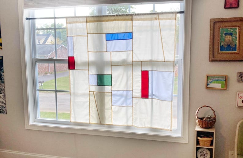 May include: A window curtain featuring a geometric grid pattern on a cream-colored background. The design includes rectangular blocks of solid blue, red, and green fabric integrated into the panel, which is suspended from a thin black rod.