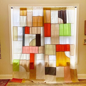 May include: A pair of curtains featuring a patchwork design composed of rectangular and square fabric panels in various colors including white, orange, yellow, lime green, brown, and tan, hanging from a rod over a window frame.
