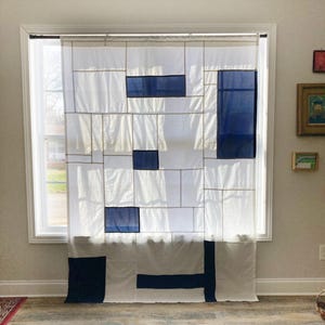 May include: A white curtain featuring a geometric grid pattern with rectangular navy blue fabric panels. The design consists of thin vertical and horizontal lines intersecting to create various rectangular sections across the semi-sheer fabric.
