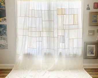 May include: A white curtain featuring a geometric grid pattern composed of thin, tan-colored lines. The fabric is semi-sheer, allowing light to pass through, and the curtain hangs from the top of the window frame, pooling slightly on the floor.