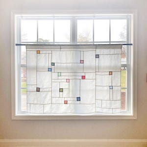 Pode incluir: Cortina de janela branca com um design geométrico. A cortina apresenta um padrão de grade com detalhes quadrados coloridos em tons de vermelho, azul, verde e castanho. A cortina está pendurada em frente a uma janela.
