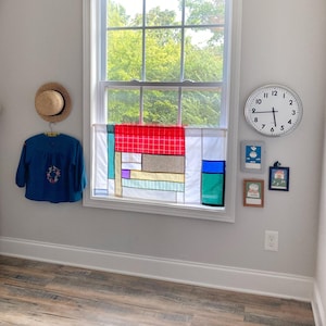 May include: A window features a patchwork curtain with red, white, blue, green, and purple geometric panels. To the left, a straw hat hangs above a blue long-sleeve shirt with floral embroidery. To the right, a round wall clock and three small framed pictures are displayed on the wall.