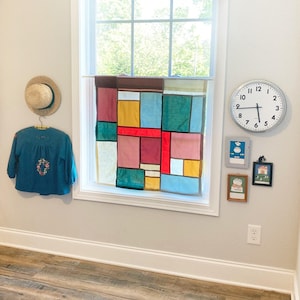 May include: A teal long-sleeve shirt with floral embroidery, a straw hat, a rectangular patchwork curtain with multi-colored fabric blocks, a round wall clock, and three small framed pictures hanging on a light-colored wall near a window.