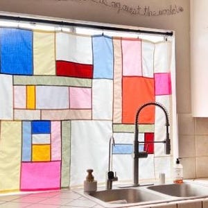 May include: A window curtain featuring a patchwork design of rectangular fabric panels in various colors including white, blue, yellow, red, pink, and light green, separated by thin dark lines, hanging over a kitchen sink with a metal faucet.