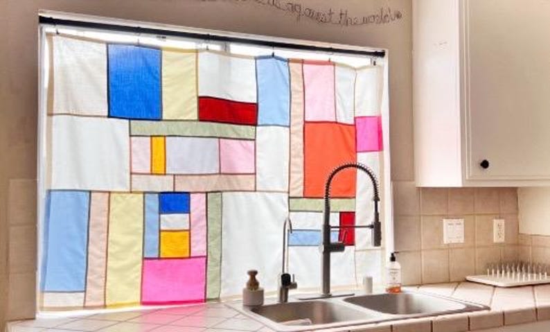 May include: A window curtain featuring a patchwork design of rectangular fabric panels in various colors including white, blue, yellow, red, pink, and light green, separated by thin dark lines, hanging over a kitchen sink with a metal faucet.