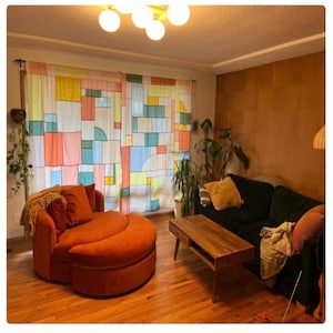 May include: A living room featuring a round orange velvet chair with a matching ottoman, a dark green sofa with a yellow throw pillow, a wooden coffee table, and sheer curtains with a multicolored geometric block pattern covering the window.