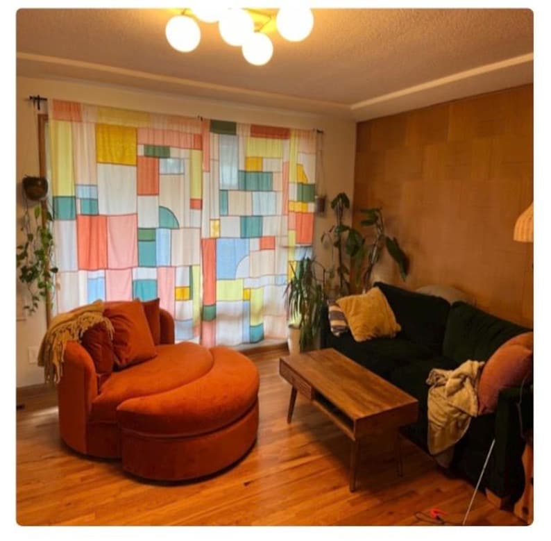 May include: A living room featuring a round orange velvet chair with a matching ottoman, a dark green sofa with a yellow throw pillow, a wooden coffee table, and sheer curtains with a multicolored geometric block pattern covering the window.