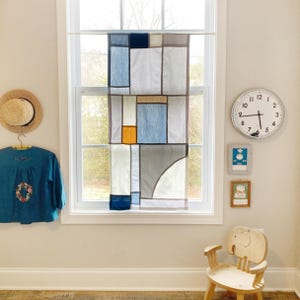 May include: A rectangular window hanging featuring a geometric patchwork design with various fabric panels in shades of blue, gray, white, and a single mustard yellow square, suspended from a thin horizontal rod against a window pane.