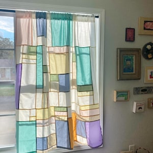 May include: A window curtain featuring a patchwork design of rectangular fabric panels in shades of white, light blue, teal, yellow, orange, and purple, separated by thin dark lines, hanging from a rod over a window frame.