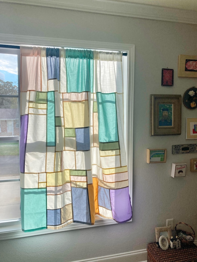 May include: A window curtain featuring a patchwork design of rectangular fabric panels in shades of white, light blue, teal, yellow, orange, and purple, separated by thin dark lines, hanging from a rod over a window frame.