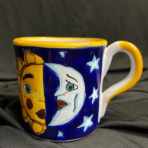 May include: A ceramic mug with a celestial design. The mug features a yellow sun and a white moon on a dark blue background with white stars. The handle and rim are yellow. The mug is approximately 4 inches tall.
