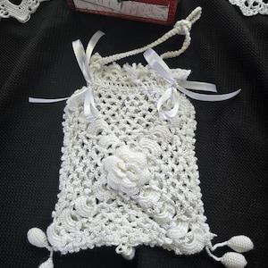 Pode incluir: Uma bolsa de crochê branca feita à mão com fecho de cordão e fitas de cetim brancas. A bolsa apresenta um design floral e elementos decorativos de crochê. A bolsa tem aproximadamente 15 cm de altura e 10 cm de largura.