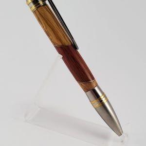 Op de afbeelding: Een handgemaakte pen met een houten vat met afwisselende secties van licht en donker hout. De pen heeft een zilverkleurige punt en accenten, met goudkleurige banden. Een zwarte clip is aan de pen bevestigd.