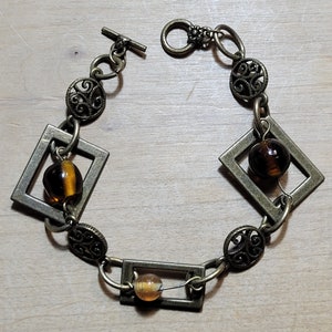 May include: A bronze bracelet with a chain link design and three amber glass beads. The bracelet has square and circular decorative elements.