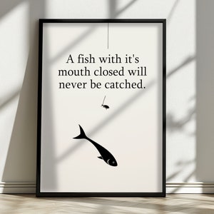 Op de afbeelding: Ingelijste kunstdruk met een minimalistisch ontwerp. Het kunstwerk toont een zwarte visillustratie met een vishaak en de tekst: "A fish with it's mouth closed will never be catched."