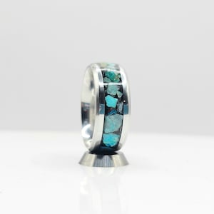 May include: A silver ring with a band of turquoise stones set in a black resin. The ring is on a silver stand.