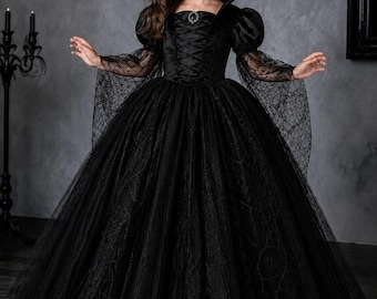 May include: A floor-length black gown featuring a high, stiff collar, puff sleeves, and long, sheer lace sleeves with a spiderweb pattern. The bodice has a lace-up front detail and a central brooch, while the skirt is made of layered tulle.