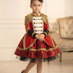 May include: A red and white ringmaster costume featuring a jacket with gold fringe epaulets, black sequined cuffs, and a full skirt with red sequins and gold trim. The outfit includes a small black top hat with gold and red decorative accents.