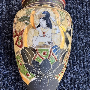 May include: A ceramic vase featuring a central figure with black hair and gold accents, seated above a large black lotus flower. The design includes intricate gold, green, and red detailing, with additional figures on the sides and a deep red rim.