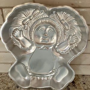 May include: A vintage aluminum cake pan in the shape of a doll. The pan features a detailed embossed design of a smiling baby with bows in its hair and outstretched arms. The pan is silver and appears to be unused.