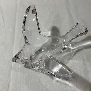 May include: A clear glass bird-shaped decorative bowl. The bowl has a wide, open design with outstretched wings and a curved tail. The glass is smooth and transparent, allowing light to pass through. The bowl sits on a white surface.