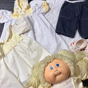 May include: A doll with blonde yarn hair and blue eyes wearing a yellow dress, accompanied by a white dress with yellow accents, a white bonnet, dark blue denim shorts, and a pair of white shoes with red stripes, all arranged together.