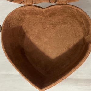 May include: A heart-shaped, brown, suede-like box with a matching lid and a thin, brown strap. The interior of the box is also brown. The box is open, revealing the inside. The box is sitting on a white surface.