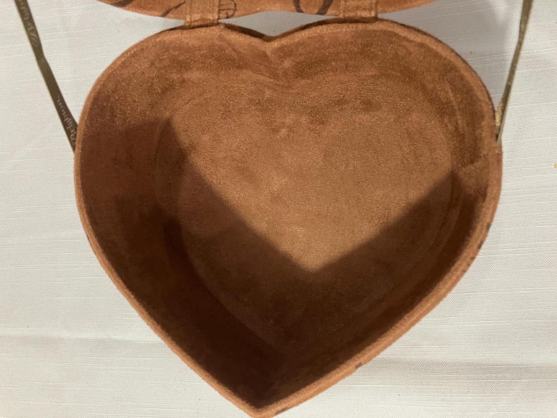 May include: A heart-shaped, brown, suede-like box with a matching lid and a thin, brown strap. The interior of the box is also brown. The box is open, revealing the inside. The box is sitting on a white surface.