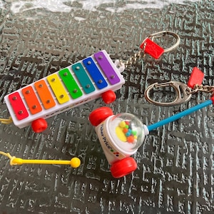 May include: Two miniature toy keychains featuring a rainbow-colored xylophone with a yellow mallet on a string and a white Fisher-Price corn popper toy with red wheels and a clear dome containing colorful balls, both attached to silver metal key rings.