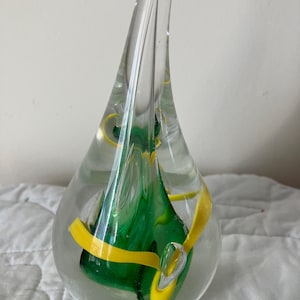 May include: A clear glass art piece with a teardrop shape. The interior features swirls of green and yellow glass. The top of the piece has a loop. The glass is smooth and the colors are vibrant.