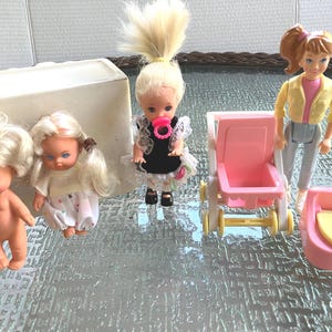May include: A collection of three small dolls, a pink plastic stroller, and a pink potty chair. One doll is nude, one wears a white patterned dress, and one has blonde hair in a high ponytail with a black dress, white lace, and a pink pacifier.