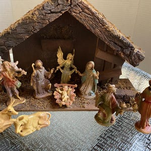 May include: A nativity scene set featuring a wooden stable with a bark-covered roof and moss accents. The set includes figurines of a central infant, two adults, an angel, three figures in robes, and two cattle, all arranged on a wooden base.