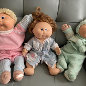 May include: Three Cabbage Patch Kids dolls with yarn hair and blue eyes. The left doll wears a pink top and blue-and-white striped leggings. The center doll has brown hair and a blue-and-white robe. The right doll wears a light green knit outfit.