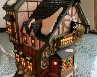 May include: A ceramic miniature building featuring a Tudor-style design with a dark shingled roof, white snow accents, and illuminated windows. A sign reads The Pied Bull, another displays 1786, and a hanging banner features the letter Q.
