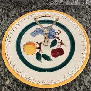 May include: A white ceramic serving tray with a yellow rim and a scalloped edge. The tray features a hand-painted design of fruit, including grapes, peaches, and cherries. The tray has a gold-tone handle.