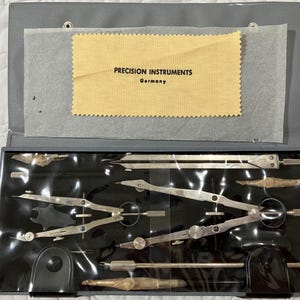 May include: A set of precision instruments in a gray case. The case is open, revealing various silver-colored drafting tools. A yellow cloth with the text "PRECISION INSTRUMENTS Germany" is visible. The tools are arranged on a black surface.