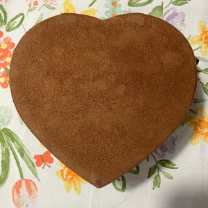 May include: A heart-shaped, brown suede box. The box is a solid color and has a soft, textured appearance. The box is sitting on a white floral patterned tablecloth.