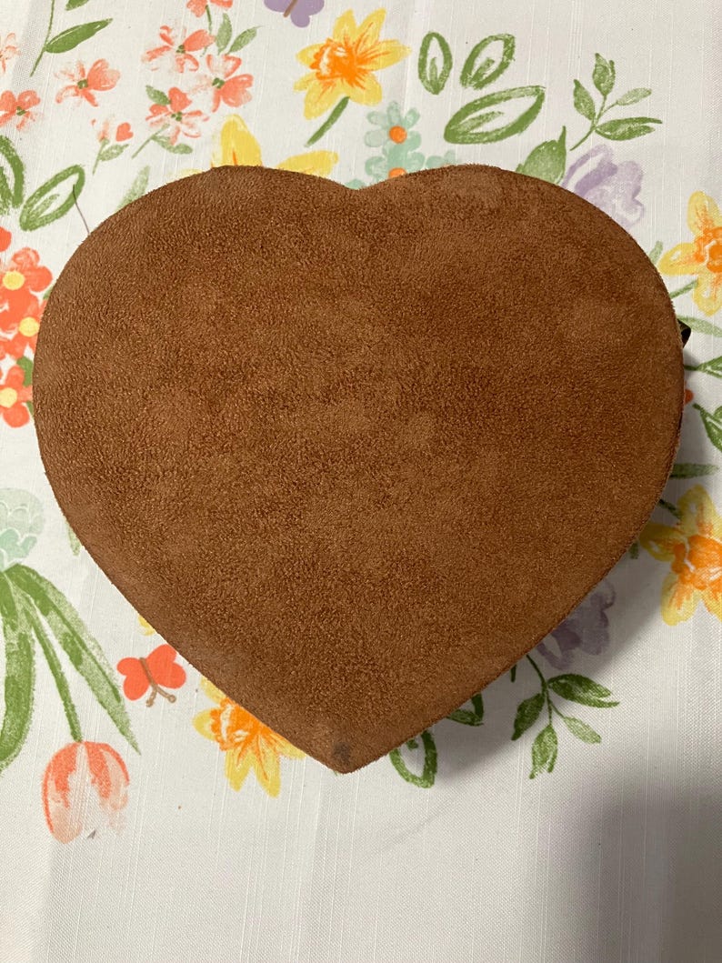 May include: A heart-shaped, brown suede box. The box is a solid color and has a soft, textured appearance. The box is sitting on a white floral patterned tablecloth.