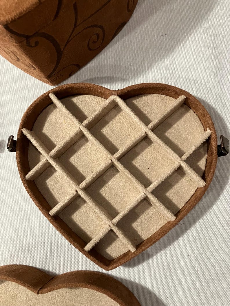 May include: A heart-shaped jewelry box with a brown exterior and a beige interior divided into compartments. The box is open, revealing the interior design. Another box is partially visible in the background.
