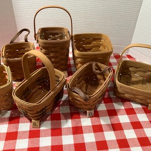 May include: A collection of six woven wooden baskets with handles, each labeled with a small wooden letter tile. The baskets vary in shape and size, featuring a light tan finish and a classic interlaced construction, arranged on a red and white checkered surface.
