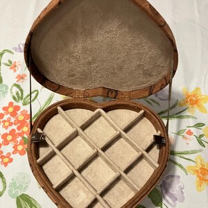 May include: Open heart-shaped jewelry box with a brown exterior and a tan interior. The box is divided into compartments and has a patterned design on the outside. The box is open, revealing the interior compartments.