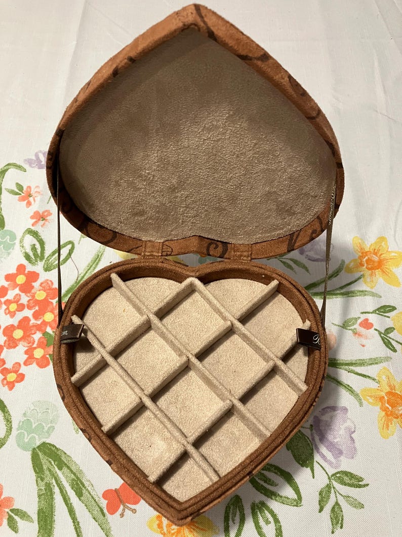 May include: Open heart-shaped jewelry box with a brown exterior and a tan interior. The box is divided into compartments and has a patterned design on the outside. The box is open, revealing the interior compartments.
