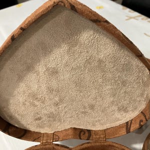 May include: Open heart-shaped jewelry box with a light brown suede interior and a brown exterior. The exterior has a darker brown decorative pattern. The box is open, revealing the soft interior. A silver ribbon is visible.