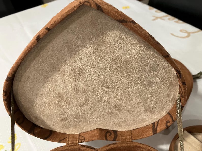 May include: Open heart-shaped jewelry box with a light brown suede interior and a brown exterior. The exterior has a darker brown decorative pattern. The box is open, revealing the soft interior. A silver ribbon is visible.