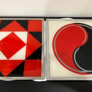 May include: Two framed abstract art pieces. One features a red, white, and black geometric pattern. The other displays a red and black yin-yang design with a silver outline, set against a cream background. Both are in silver frames.